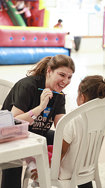 A smiling woman in a black "GLOBAL GIVING" t-shirt paints a child's face at an event, with a colorful bounce house behind them.