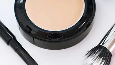Open, round, black makeup compact with light foundation, flanked by a black eyeliner pencil and a makeup brush.