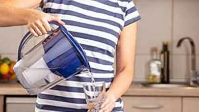 A person in a striped shirt pours water from a blue pitcher into a clear glass in a kitchen.