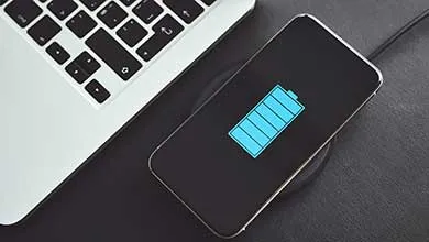 Top-down close-up of a smartphone on a wireless charging pad, displaying a large blue battery icon nearly full, next to a laptop corner on a dark surface.