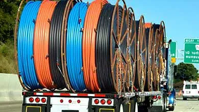 A flatbed semi-truck carries large black, orange, and blue utility cable spools on a highway under a clear blue sky. An overhead green sign shows Exit 152B for MacArthur Drive and Pike Avenue in half a mile. A white van and a yellow construction vehicle are in adjacent lanes.