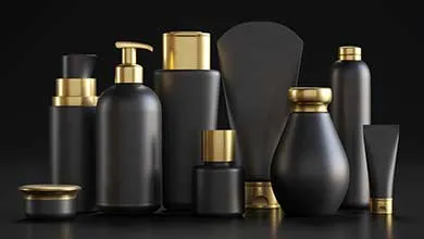Matte black cosmetic and skincare containers with gold caps, pump dispensers, and metallic accents, including cylindrical bottles, a round vessel, a small jar, and a tube, arranged on a dark background.