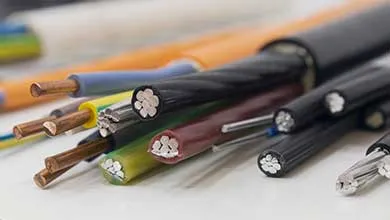 Close-up of several electrical cables with cut ends, revealing copper and aluminum wires inside. The cables are black, blue, yellow, green, and brown, overlapping on a white surface.